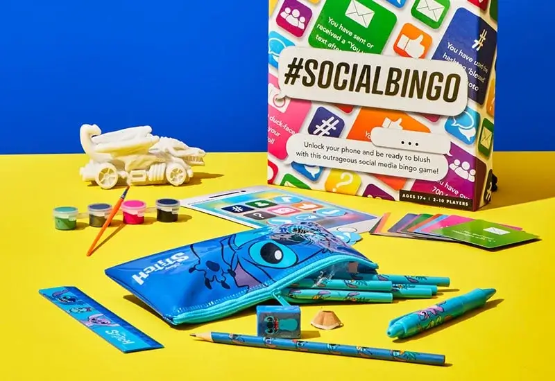 Colorful assortment of items on a yellow surface with a blue background, featuring a '#SOCIALBINGO' game box with social media icons, surrounded by art supplies, a Stitch pencil case, a toy car, and bingo game cards.